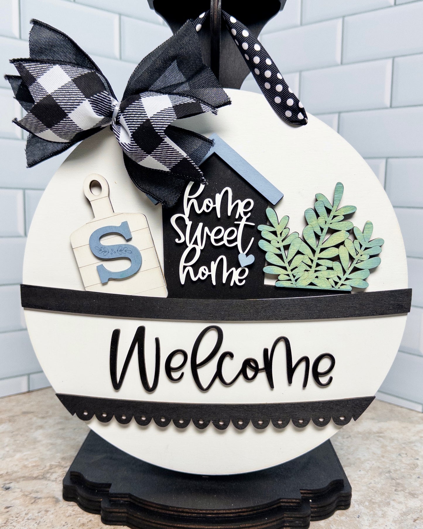 Decorative sign with 'Home Sweet Home' and 'Welcome' text, featuring a black and white checkered bow.