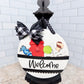 Decorative doorbell cover with colorful buttons and 'Welcome' text, featuring a black and white checkered bow, against a white brick wall background.