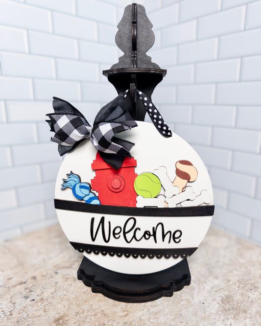 Decorative doorbell cover with colorful buttons and 'Welcome' text, featuring a black and white checkered bow, against a white brick wall background.