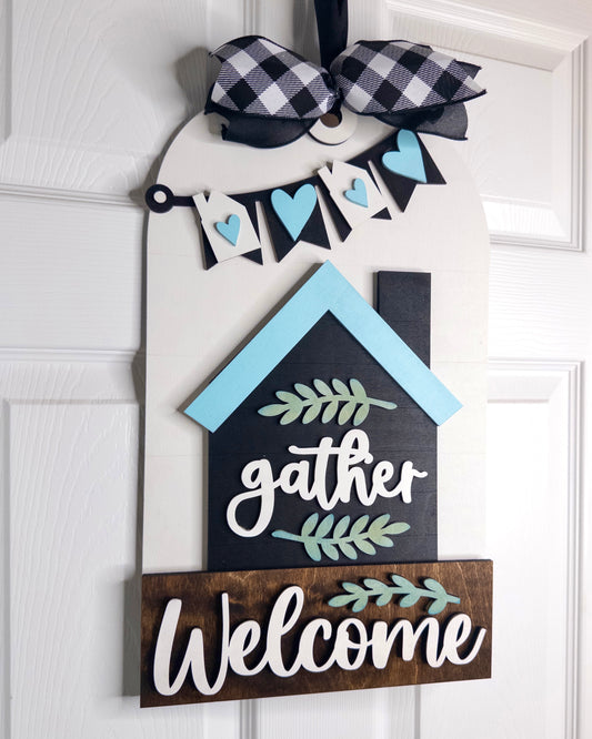 Decorative door hanger with 'gather' and 'Welcome' text on a white door.
