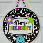 Decorative round sign with 'Mrs. BILBREY' text, colorful characters, and a black and white pattern on a white door.