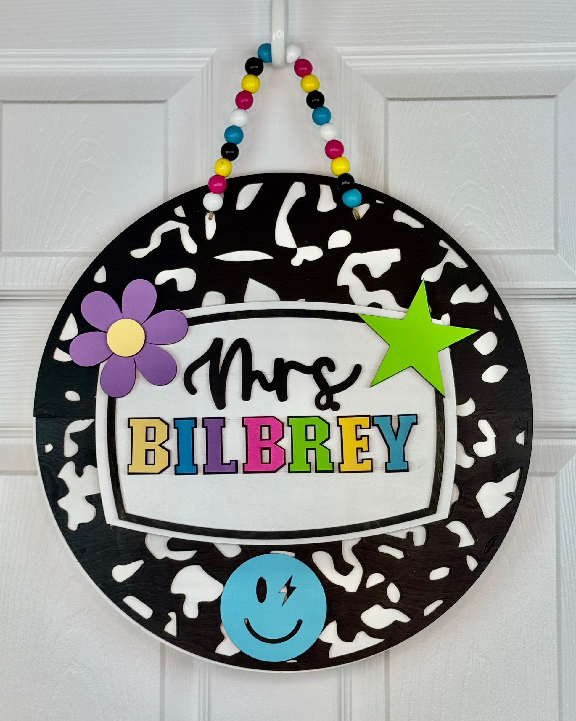 Decorative round sign with 'Mrs. BILBREY' text, colorful characters, and a black and white pattern on a white door.