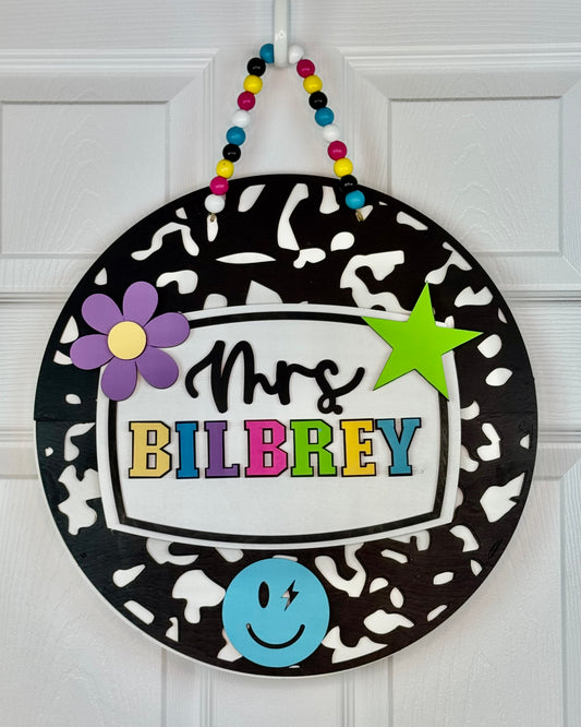 Decorative round sign with 'Mrs. BILBREY' text, colorful characters, and a black and white pattern on a white door.